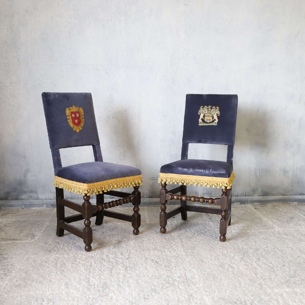 A matched pair of dark oak side chairs upholstered in dark blue velvet, featuring embroidered heraldic crests on the backrests and gold tassel fringe around the seat borders. View XII