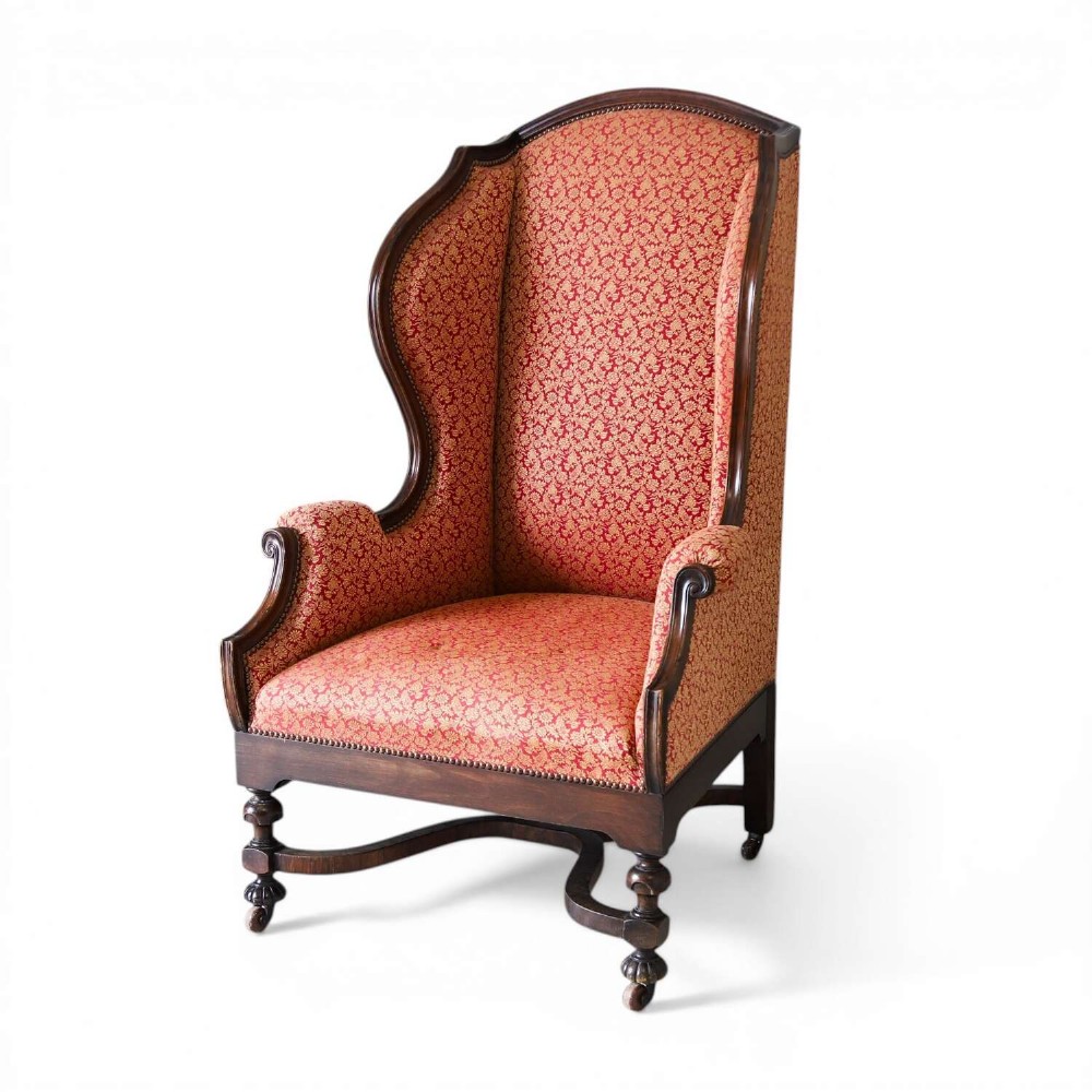 A mid-19th-century high-back wing armchair upholstered in red and gold patterned fabric with carved dark timber legs on casters.