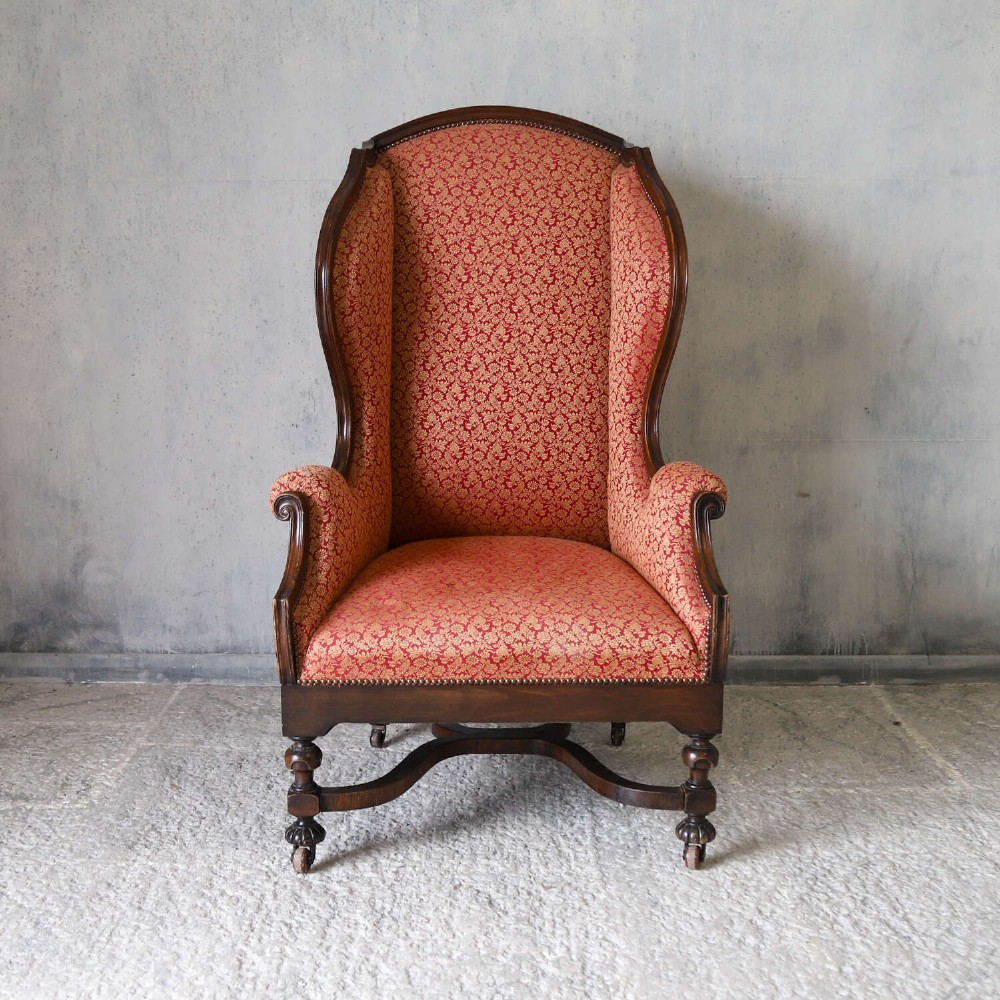 A mid-19th-century high-back wing armchair upholstered in red and gold patterned fabric with carved dark timber legs on casters. View II
