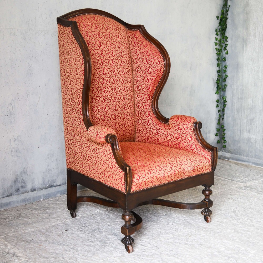 A mid-19th-century high-back wing armchair upholstered in red and gold patterned fabric with carved dark timber legs on casters. View III