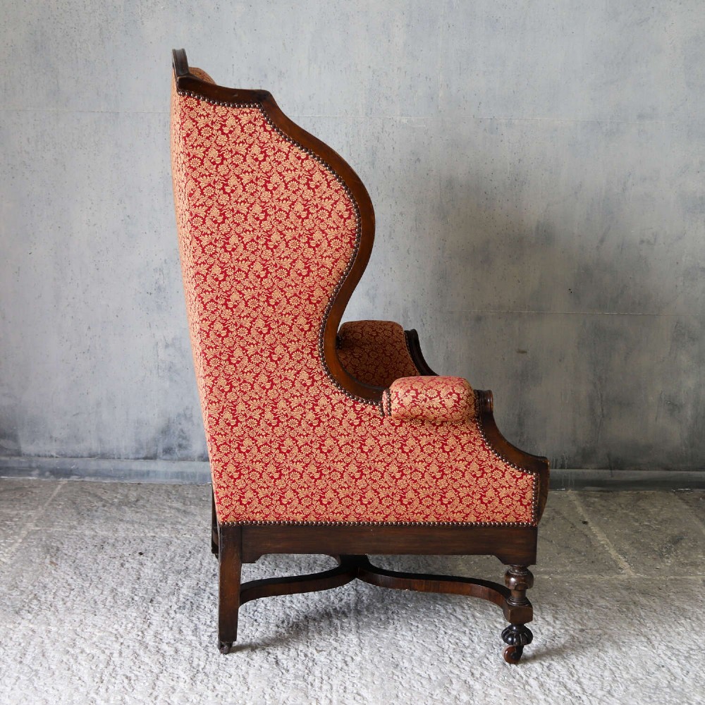 A mid-19th-century high-back wing armchair upholstered in red and gold patterned fabric with carved dark timber legs on casters. View X