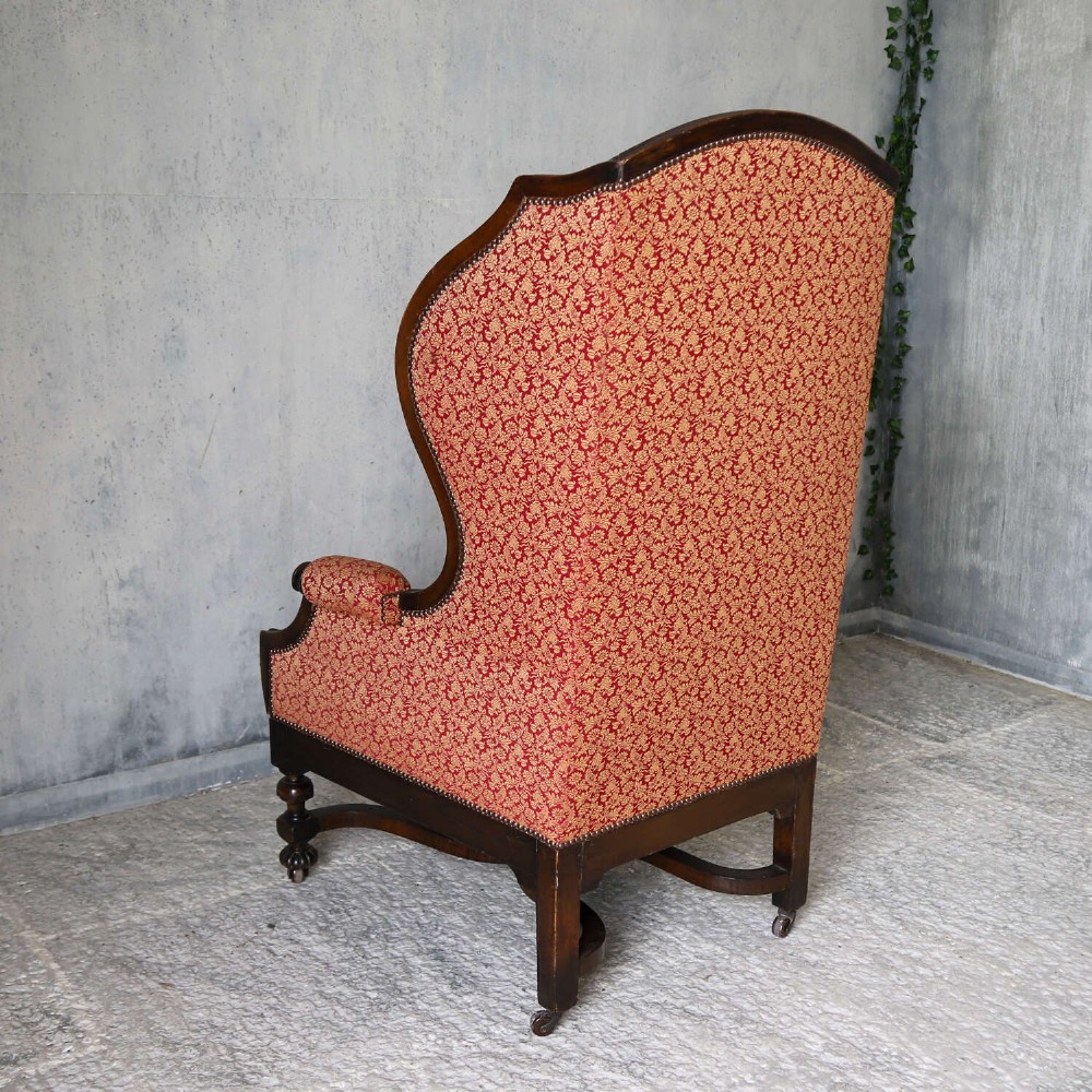 A mid-19th-century high-back wing armchair upholstered in red and gold patterned fabric with carved dark timber legs on casters. View XII