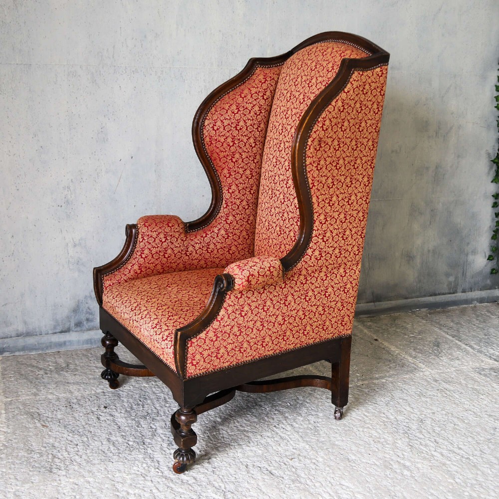 A mid-19th-century high-back wing armchair upholstered in red and gold patterned fabric with carved dark timber legs on casters. View XIII