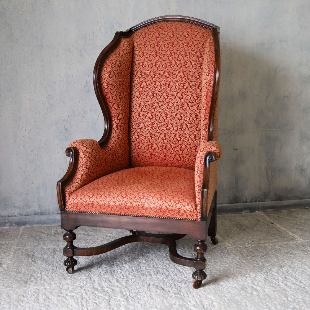A mid-19th-century high-back wing armchair upholstered in red and gold patterned fabric with carved dark timber legs on casters. View XIV