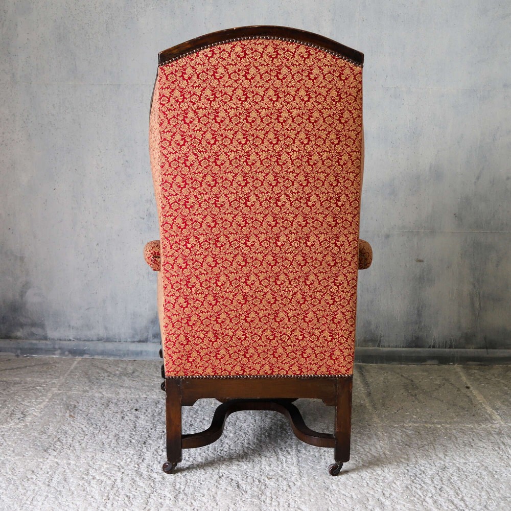 A mid-19th-century high-back wing armchair upholstered in red and gold patterned fabric with carved dark timber legs on casters. View XV