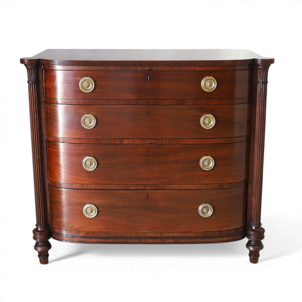 A Scottish Regency flame mahogany bowfront chest of four graduated drawers featuring brass ring handles, reeded side columns, and turned feet.