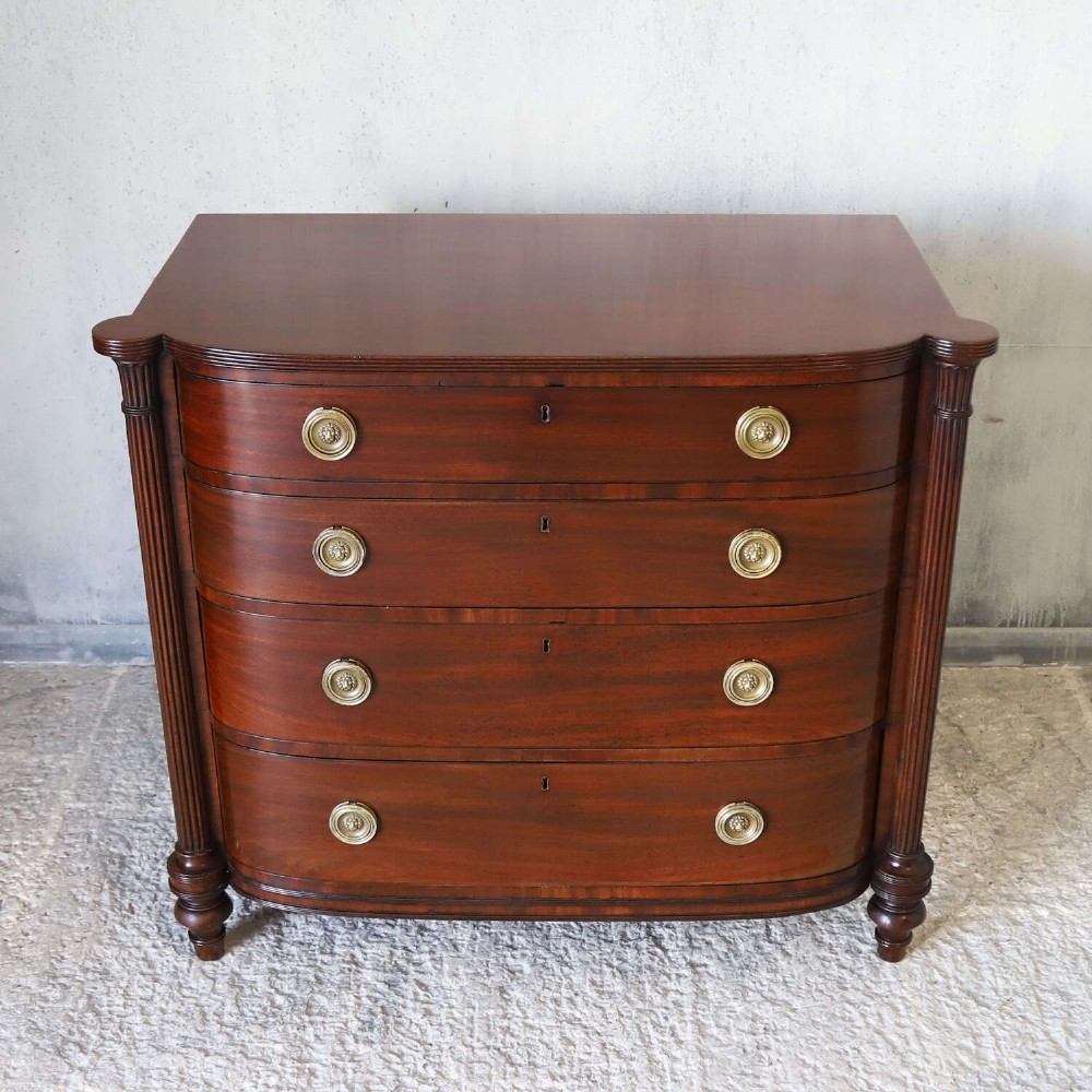 A Scottish Regency flame mahogany bowfront chest of four graduated drawers featuring brass ring handles, reeded side columns, and turned feet. View II