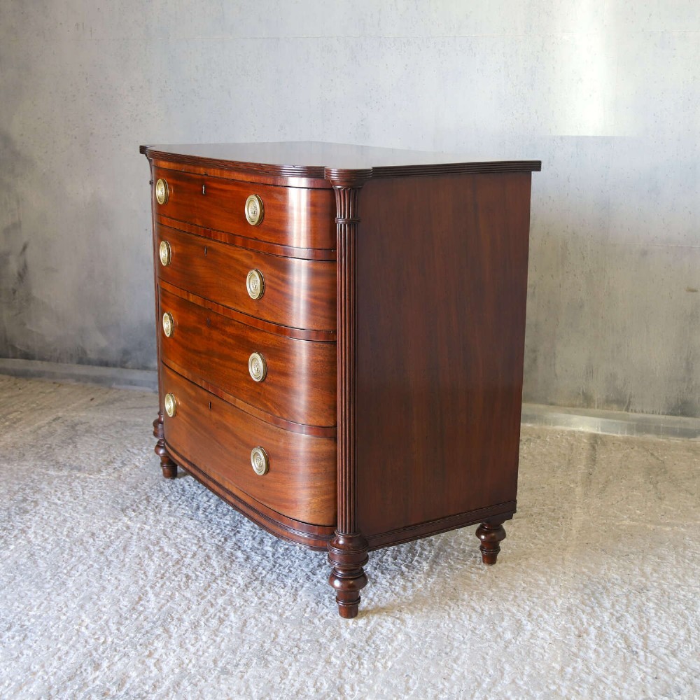 A Scottish Regency flame mahogany bowfront chest of four graduated drawers featuring brass ring handles, reeded side columns, and turned feet. View VI