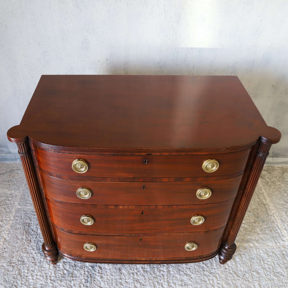 A Scottish Regency flame mahogany bowfront chest of four graduated drawers featuring brass ring handles, reeded side columns, and turned feet. View VIII