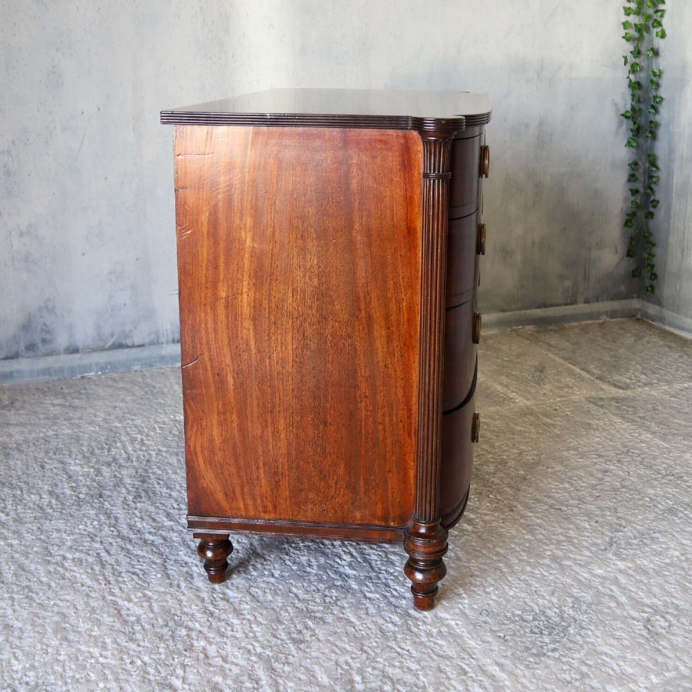 A Scottish Regency flame mahogany bowfront chest of four graduated drawers featuring brass ring handles, reeded side columns, and turned feet. View IX