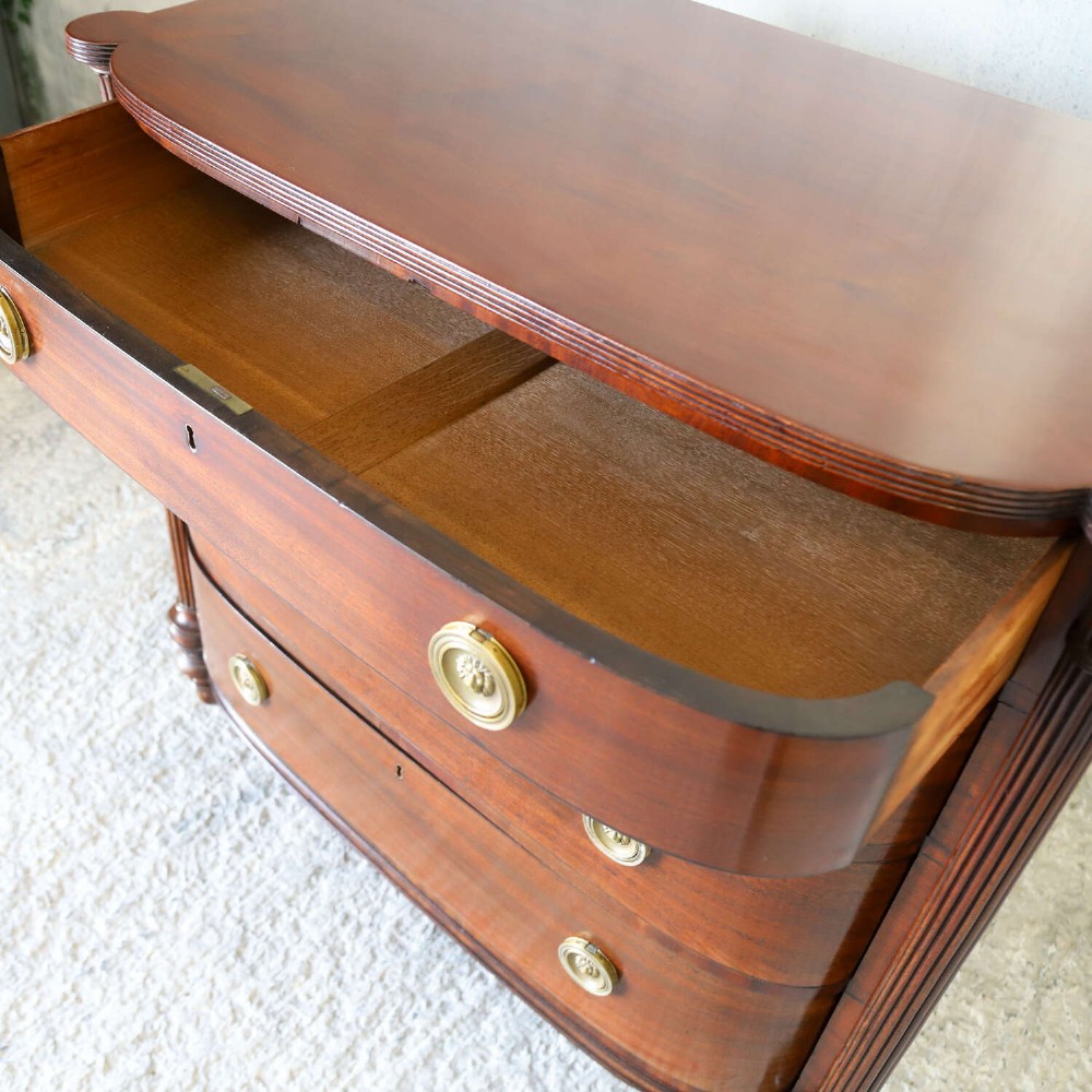 A Scottish Regency flame mahogany bowfront chest of four graduated drawers featuring brass ring handles, reeded side columns, and turned feet. View XII
