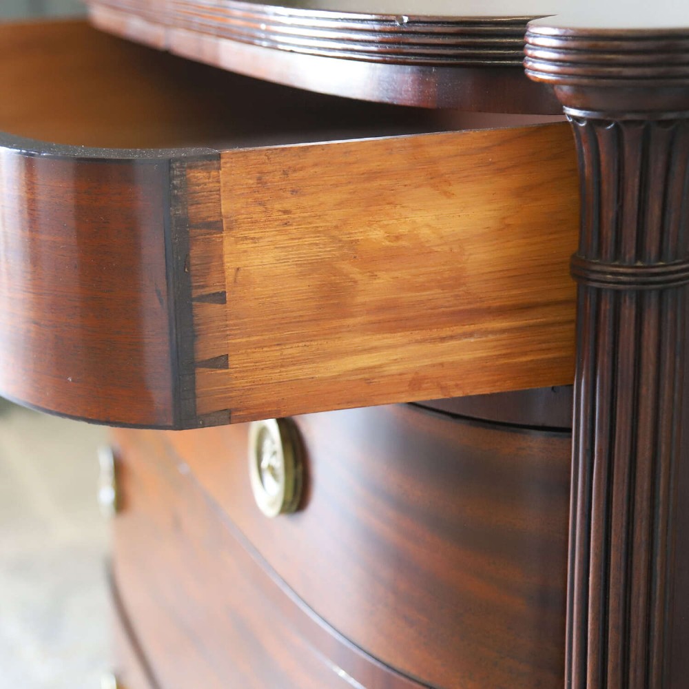 A Scottish Regency flame mahogany bowfront chest of four graduated drawers featuring brass ring handles, reeded side columns, and turned feet. View XIII