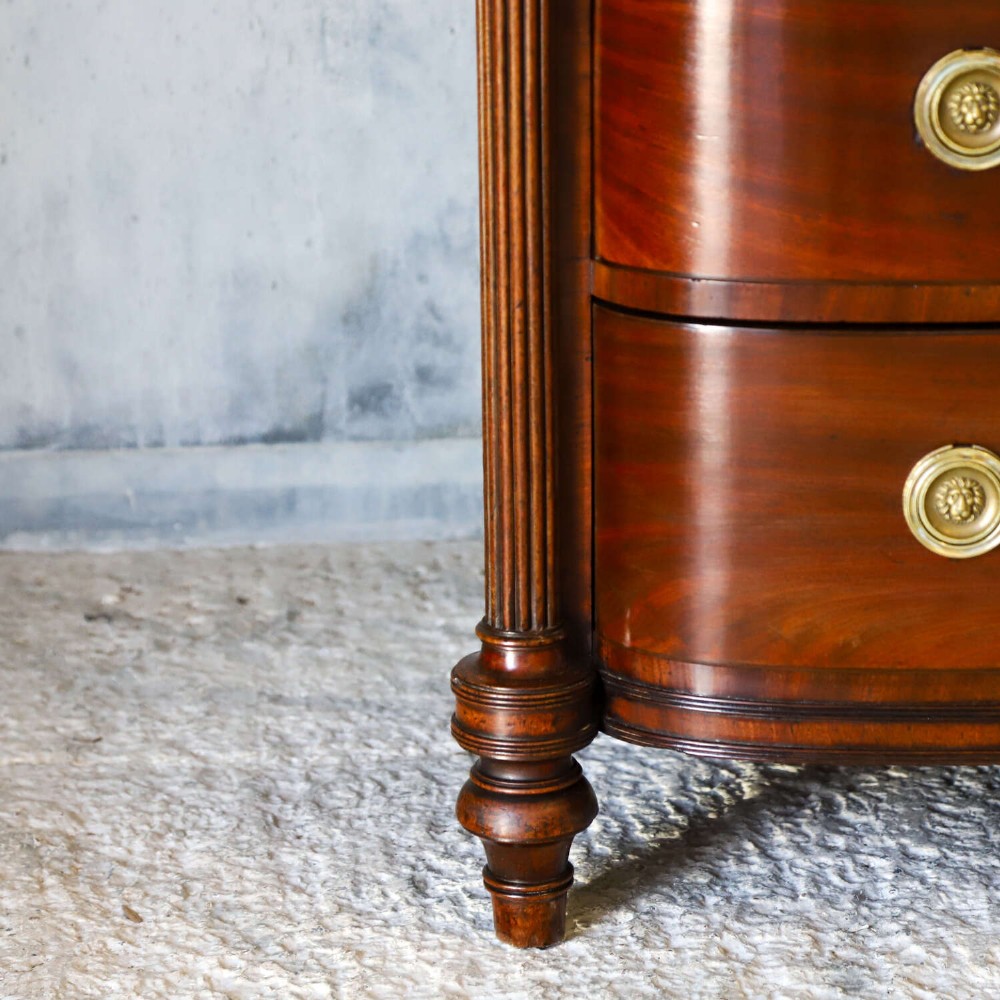 A Scottish Regency flame mahogany bowfront chest of four graduated drawers featuring brass ring handles, reeded side columns, and turned feet. View XIV