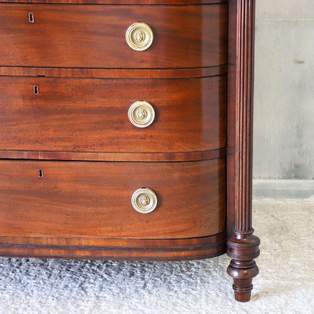 A Scottish Regency flame mahogany bowfront chest of four graduated drawers featuring brass ring handles, reeded side columns, and turned feet. View XV