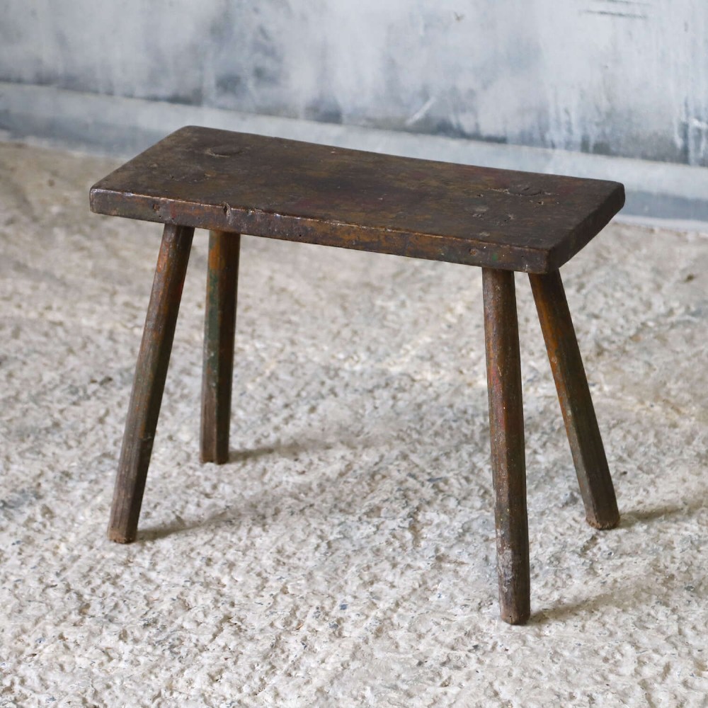 A rectangular rustic wooden milking stool with splayed cylindrical legs and a distressed dark green painted finish. View III
