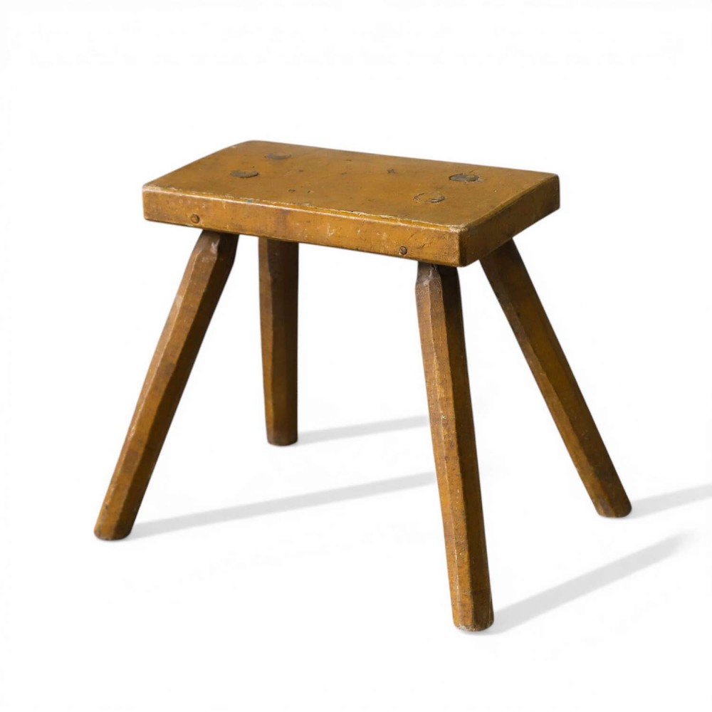 A small rustic 19th-century wooden milking stool with a rectangular top, four splayed legs, and weathered ochre yellow paint.