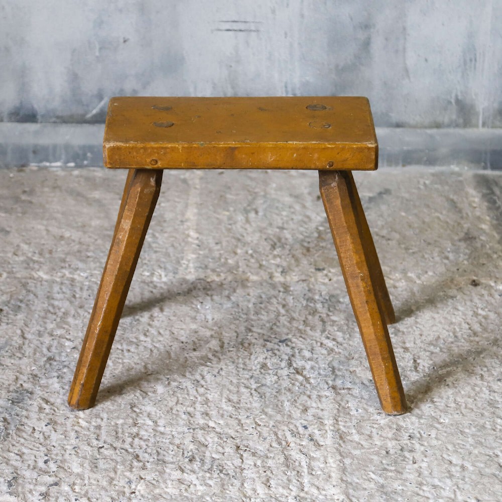 A small rustic 19th-century wooden milking stool with a rectangular top, four splayed legs, and weathered ochre yellow paint. View II