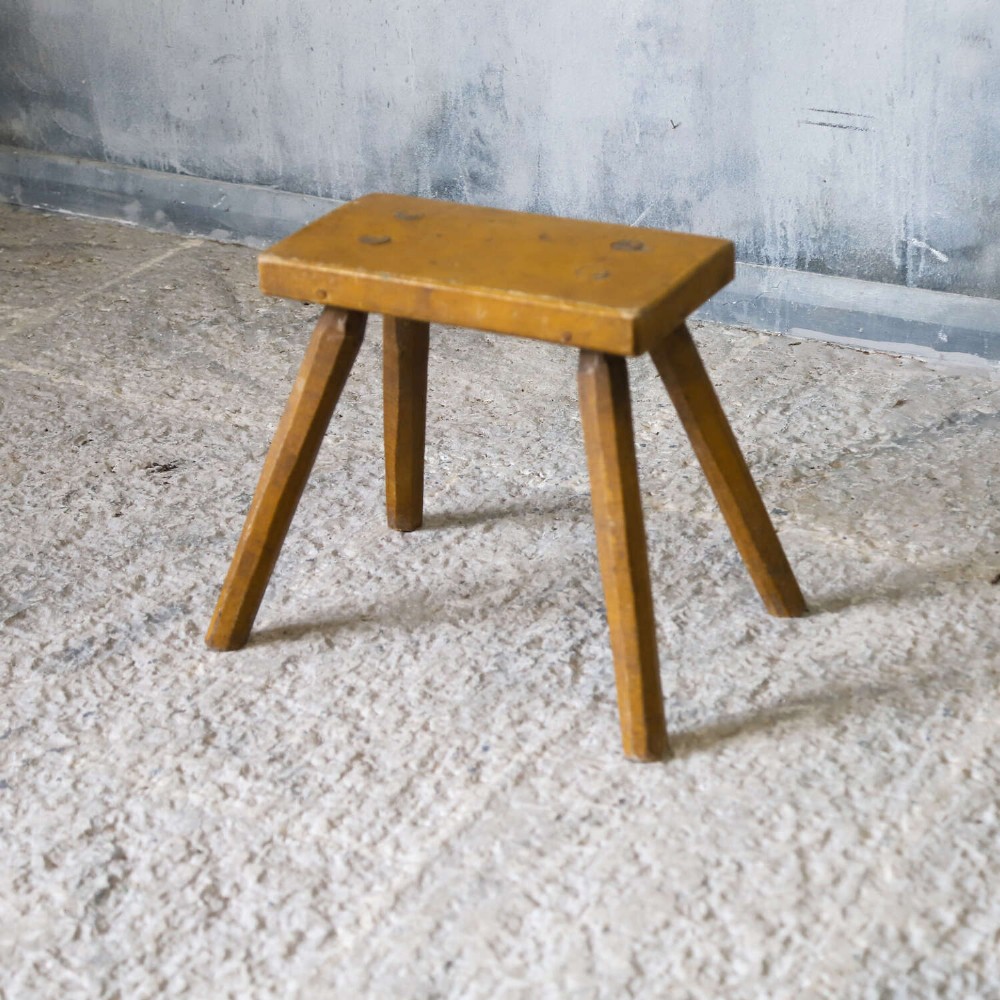 A small rustic 19th-century wooden milking stool with a rectangular top, four splayed legs, and weathered ochre yellow paint. View IV