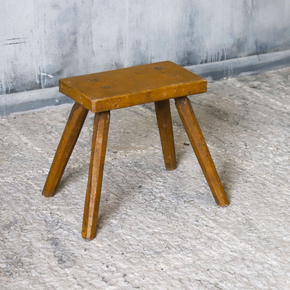 A small rustic 19th-century wooden milking stool with a rectangular top, four splayed legs, and weathered ochre yellow paint. View VIII