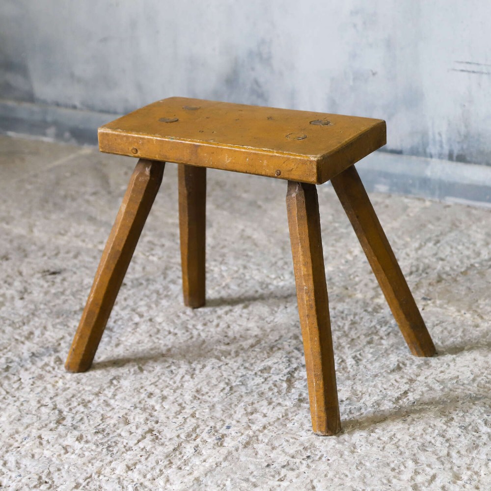 A small rustic 19th-century wooden milking stool with a rectangular top, four splayed legs, and weathered ochre yellow paint. View X