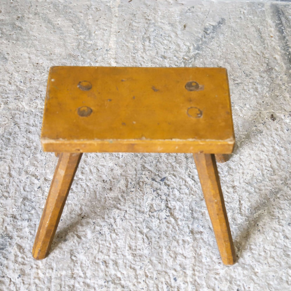 A small rustic 19th-century wooden milking stool with a rectangular top, four splayed legs, and weathered ochre yellow paint. View XI