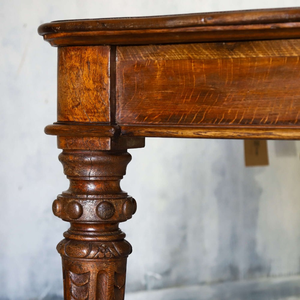 A 19th-century English oak Gothic Revival library table featuring an inset leather top and carved turned legs on brass castors. View X