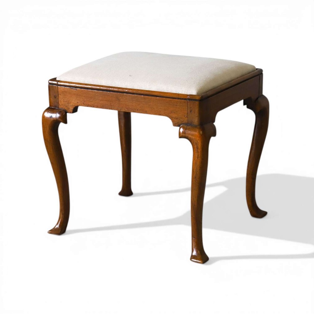An early eighteenth-century George I walnut stool featuring four shaped cabriole legs and a cream upholstered drop-in seat.