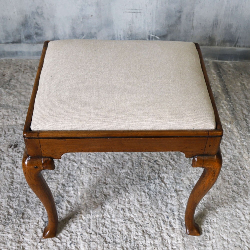 An early eighteenth-century George I walnut stool featuring four shaped cabriole legs and a cream upholstered drop-in seat. View V