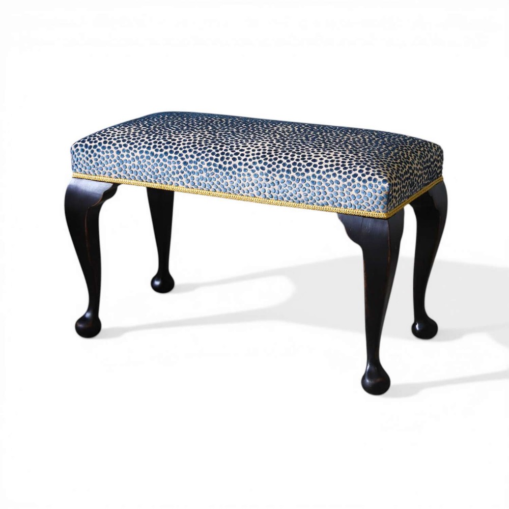 An Edwardian style ebonised wooden stool with curved cabriole legs, pad feet, gold trim braid, and a patterned blue upholstered top.