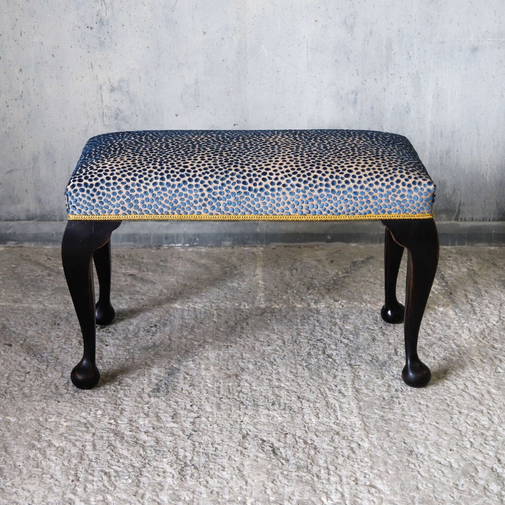 An Edwardian style ebonised wooden stool with curved cabriole legs, pad feet, gold trim braid, and a patterned blue upholstered top. View II