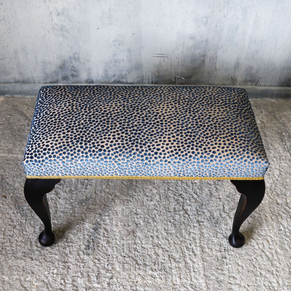 An Edwardian style ebonised wooden stool with curved cabriole legs, pad feet, gold trim braid, and a patterned blue upholstered top. View V