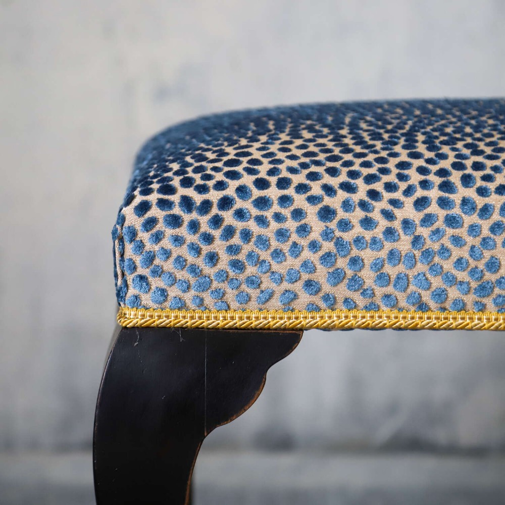 An Edwardian style ebonised wooden stool with curved cabriole legs, pad feet, gold trim braid, and a patterned blue upholstered top. View VI