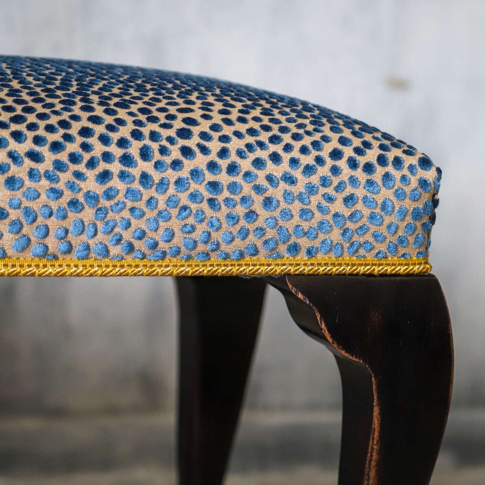 An Edwardian style ebonised wooden stool with curved cabriole legs, pad feet, gold trim braid, and a patterned blue upholstered top. View VIII