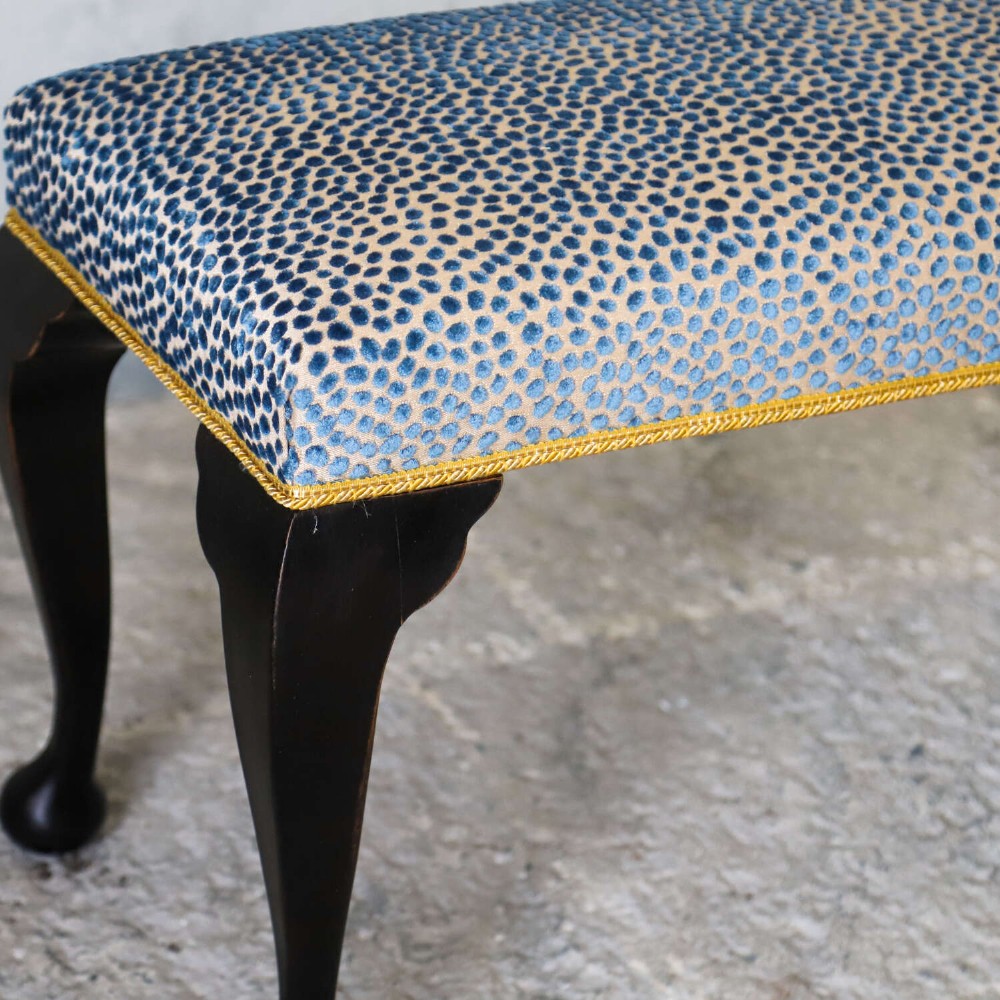An Edwardian style ebonised wooden stool with curved cabriole legs, pad feet, gold trim braid, and a patterned blue upholstered top. View IX