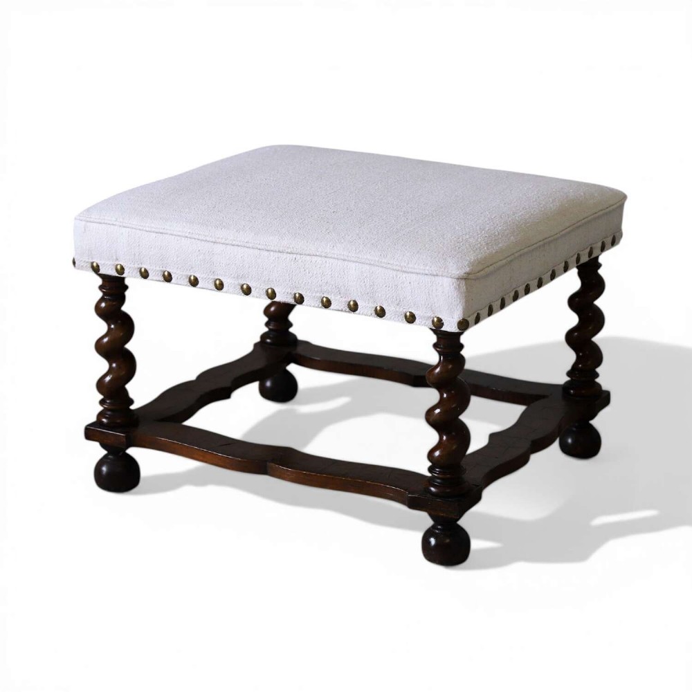 A square upholstered stool with neutral fabric and brass nailhead trim, supported by dark turned barley-twist legs and a serpentine stretcher on ball feet.
