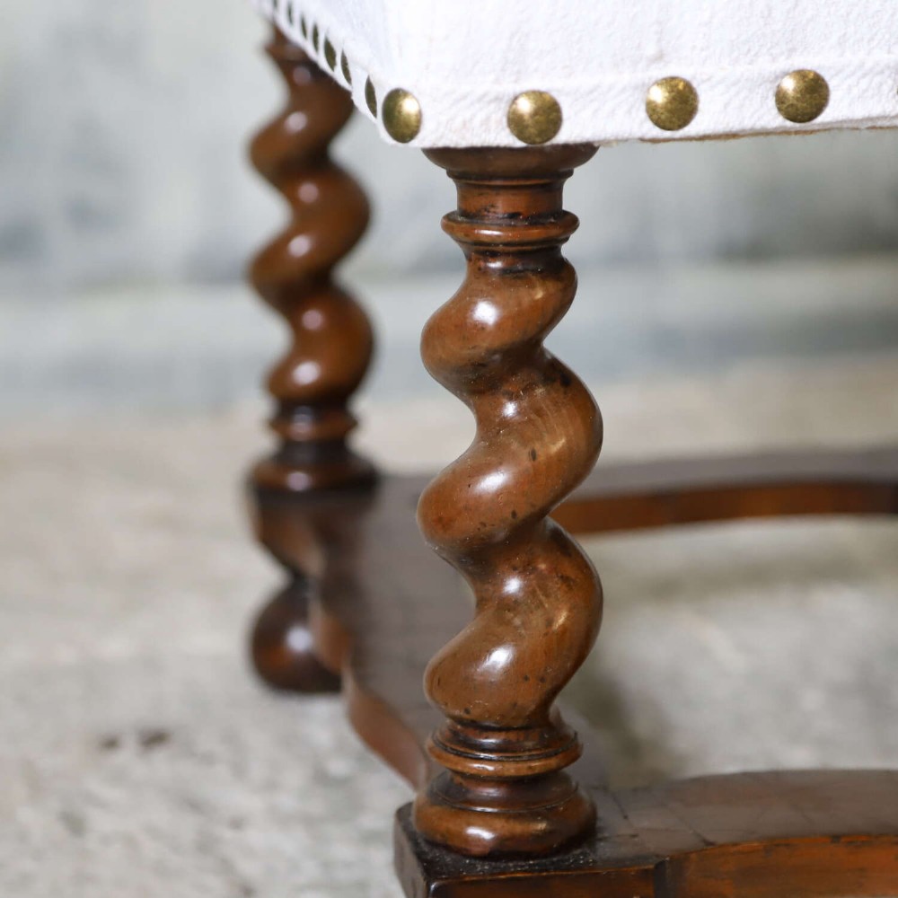 A square upholstered stool with neutral fabric and brass nailhead trim, supported by dark turned barley-twist legs and a serpentine stretcher on ball feet. View VI