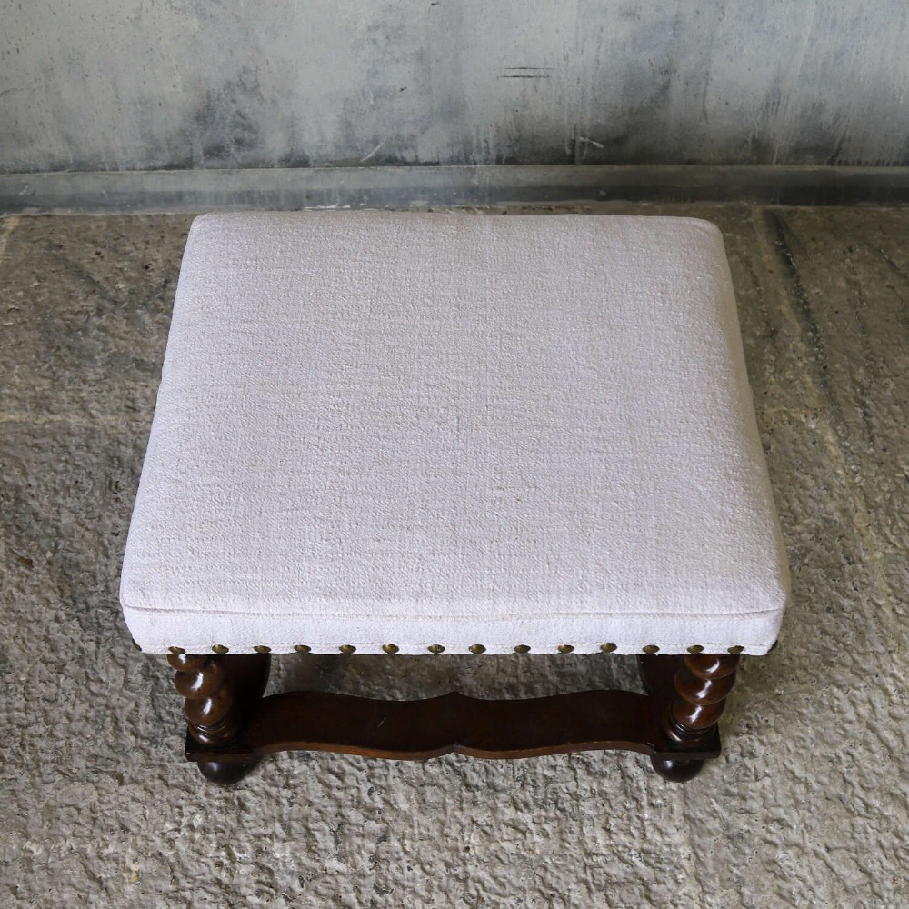 A square upholstered stool with neutral fabric and brass nailhead trim, supported by dark turned barley-twist legs and a serpentine stretcher on ball feet. View VII