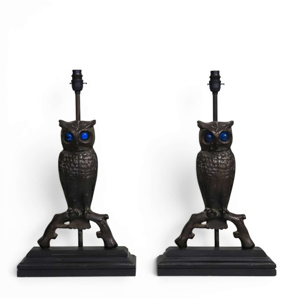 A pair of dark cast iron owl table lamps with bright blue glass eyes, perched on branch bases mounted on black plinths.