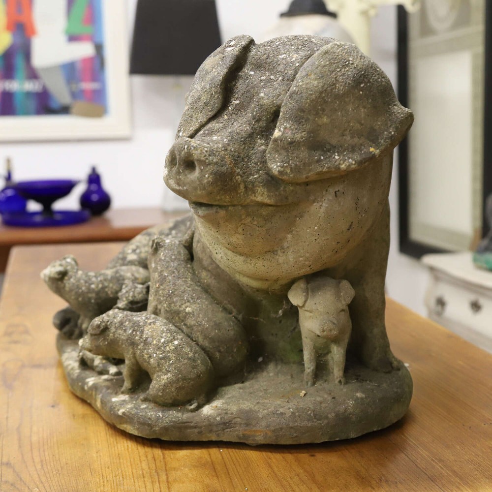 A weathered cast stone garden statue featuring a reclining sow flanked by seven small piglets on a moulded oval base. View VI