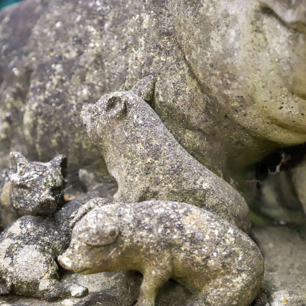 A weathered cast stone garden statue featuring a reclining sow flanked by seven small piglets on a moulded oval base. View XII