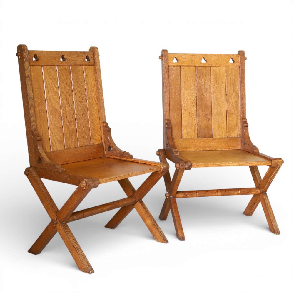 A pair of warm brown oak Glastonbury chairs featuring panelled backs with trefoil cut-outs and exposed pegged X-frame legs.