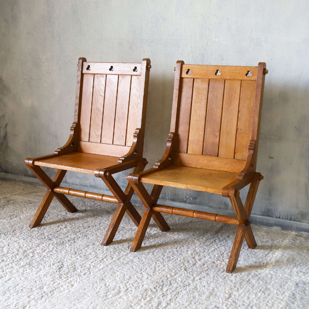 A pair of warm brown oak Glastonbury chairs featuring panelled backs with trefoil cut-outs and exposed pegged X-frame legs. View V