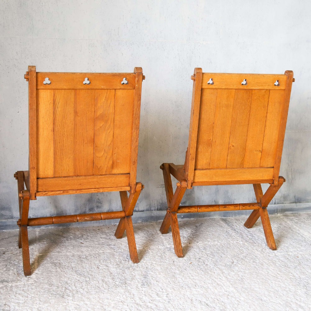A pair of warm brown oak Glastonbury chairs featuring panelled backs with trefoil cut-outs and exposed pegged X-frame legs. View X