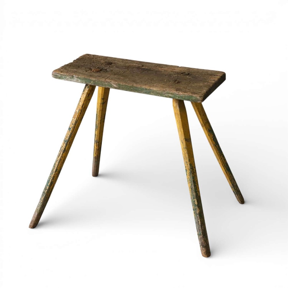 A small antique rustic timber milking stool featuring four angled splayed legs and a distressed yellow and green painted finish.