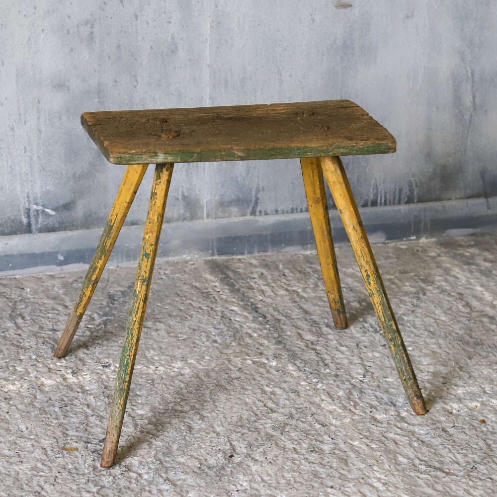 A small antique rustic timber milking stool featuring four angled splayed legs and a distressed yellow and green painted finish. View II