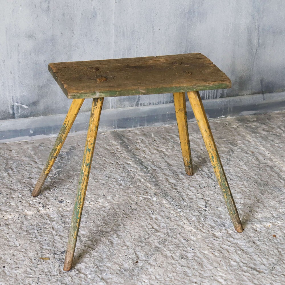 A small antique rustic timber milking stool featuring four angled splayed legs and a distressed yellow and green painted finish. View III