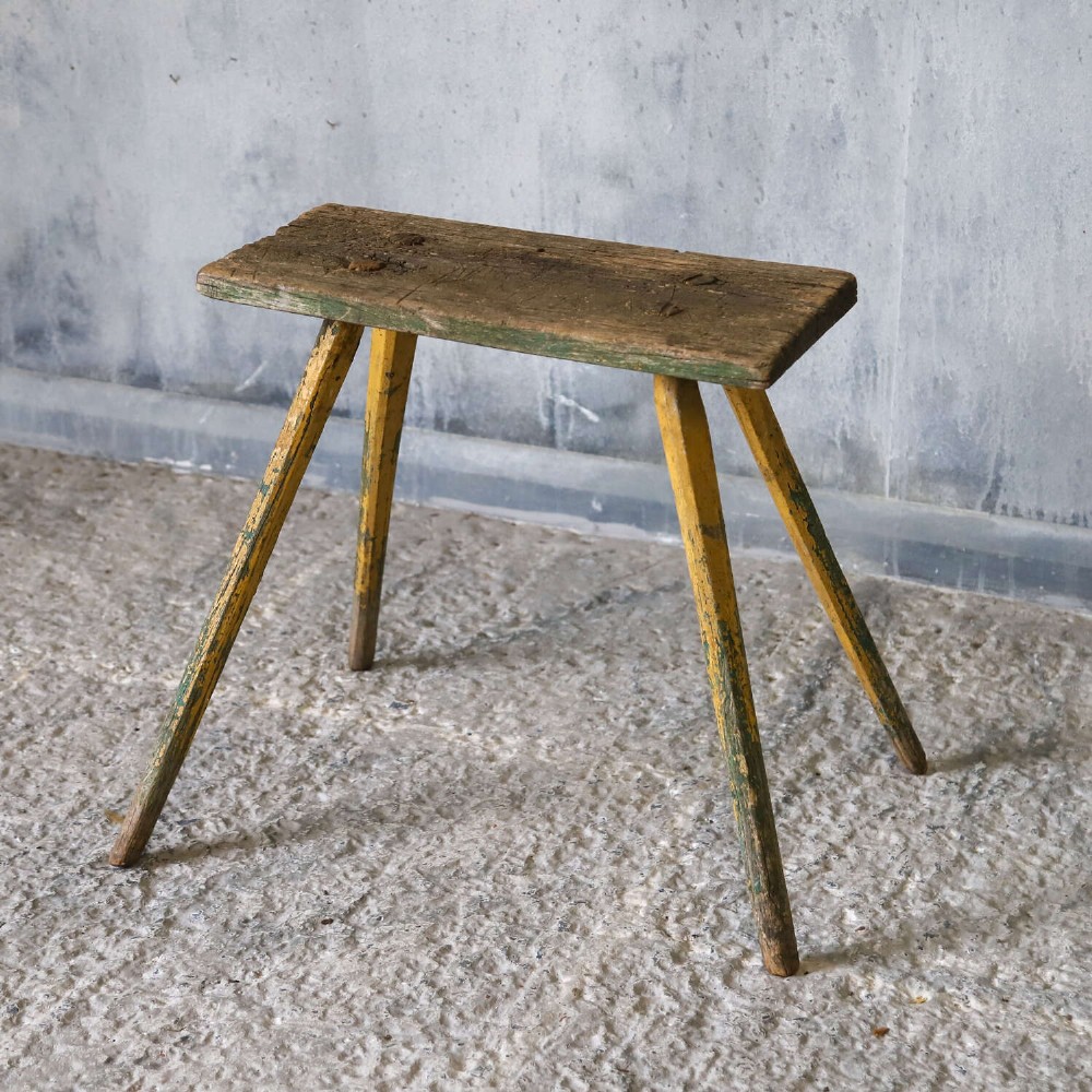 A small antique rustic timber milking stool featuring four angled splayed legs and a distressed yellow and green painted finish. View IV