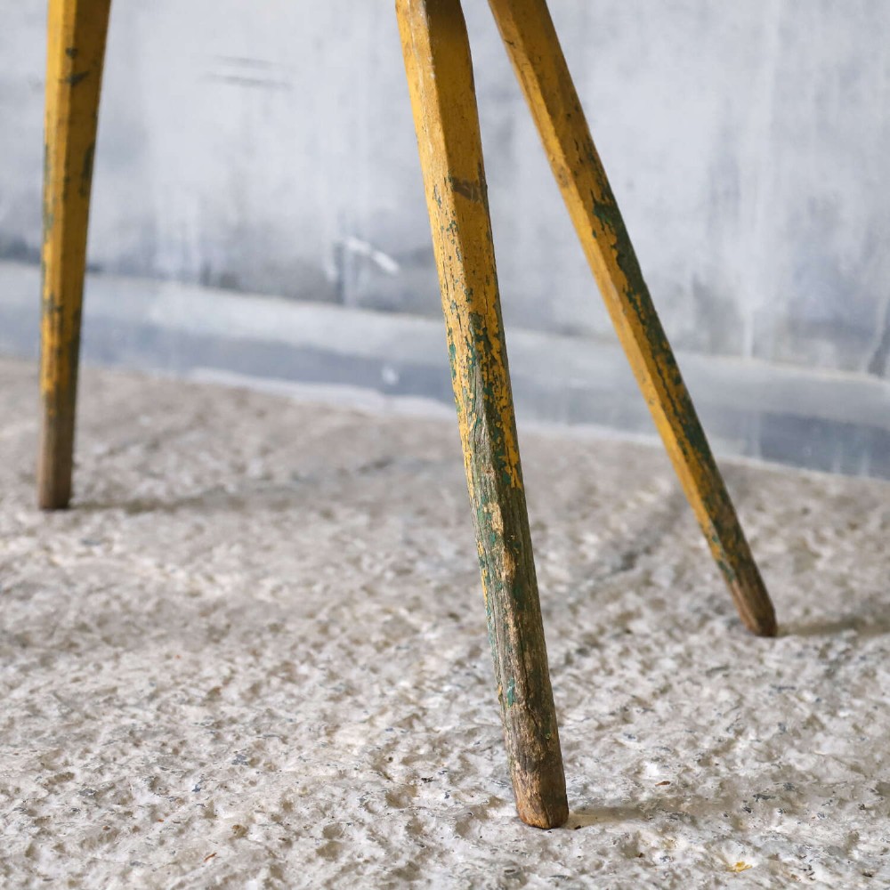 A small antique rustic timber milking stool featuring four angled splayed legs and a distressed yellow and green painted finish. View V