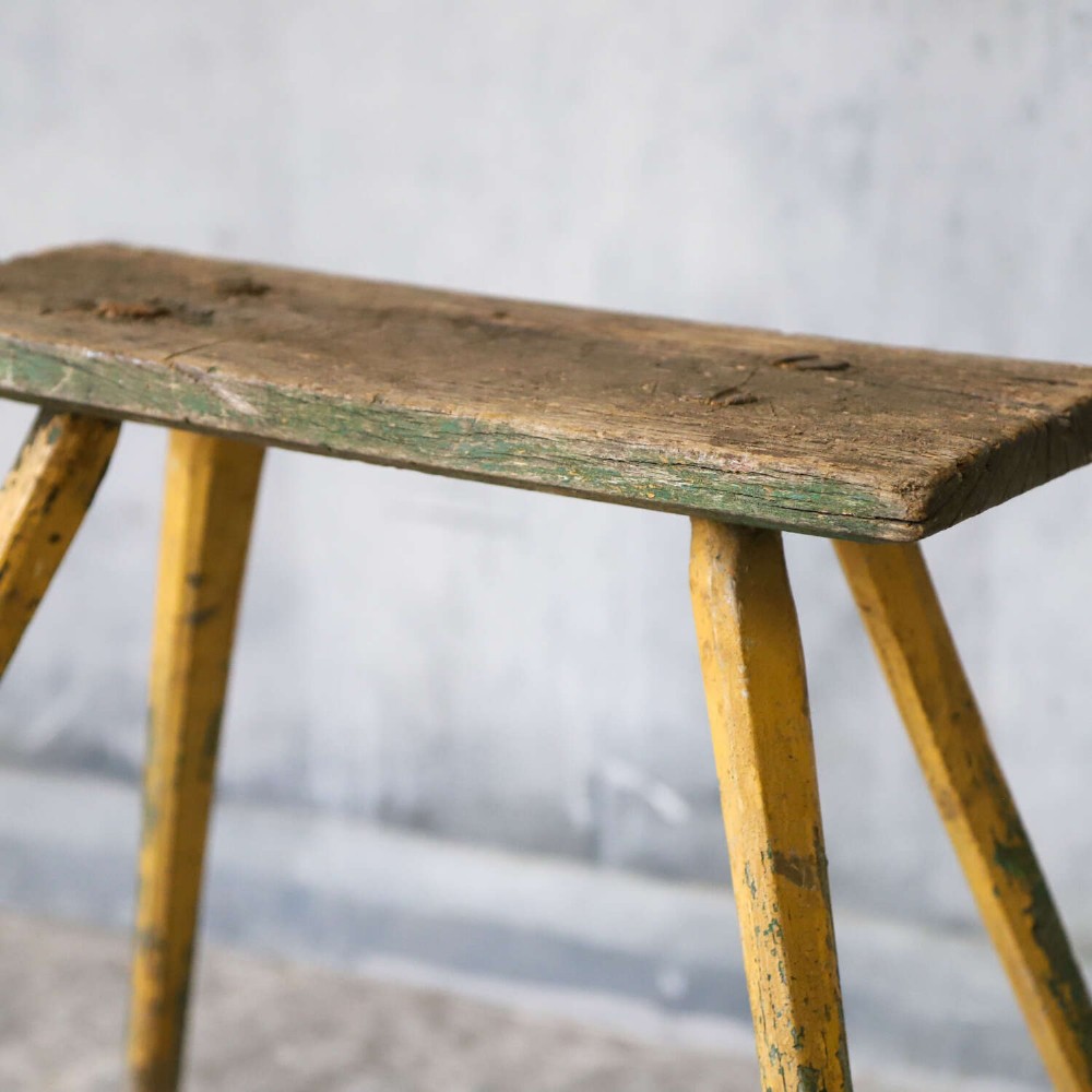 A small antique rustic timber milking stool featuring four angled splayed legs and a distressed yellow and green painted finish. View VI
