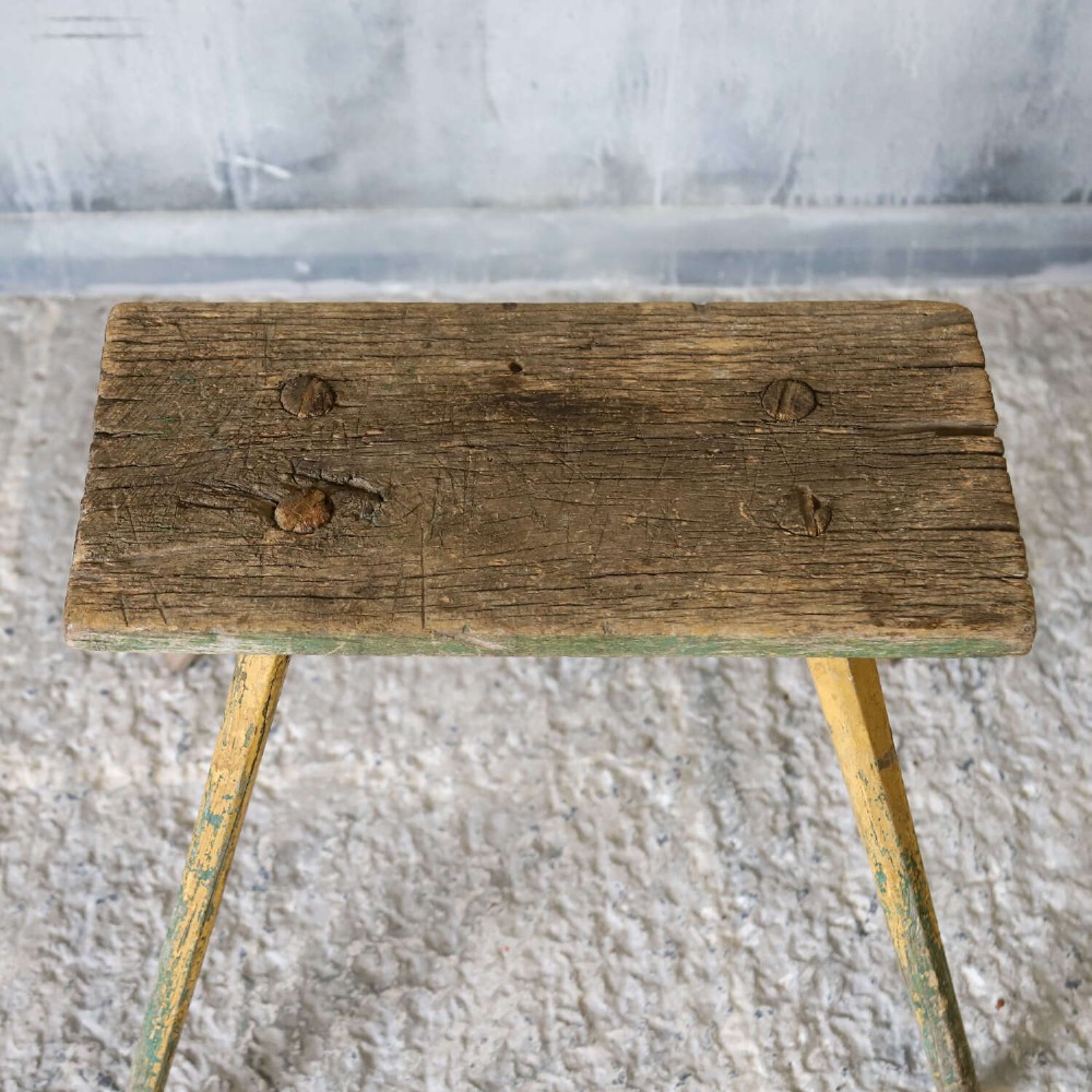 A small antique rustic timber milking stool featuring four angled splayed legs and a distressed yellow and green painted finish. View VII
